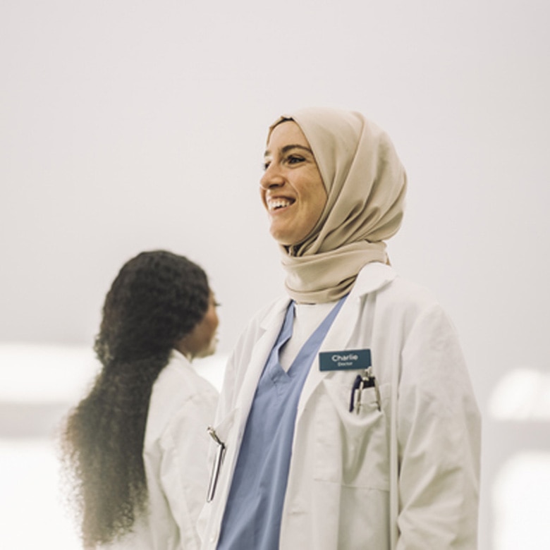 A smiling female doctor wearing a tan hijab, blue scrubs, and a white lab coat with a "Charlie" name tag.